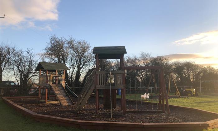 A wooden playground with climbing walls, a fireman's pole, swings, a slide and a mini tractor on grass and in front of trees.